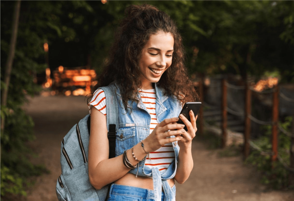 Adolescente de cabelo cacheado e castanho sorrindo para a tela do smartphone